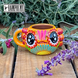 May include: Orange ceramic mug with a whimsical face design. The mug features large black eyes, a smiling mouth, and colorful floral patterns in red, pink, and turquoise. The handle is curved, and the mug is set against a wooden surface with lavender.