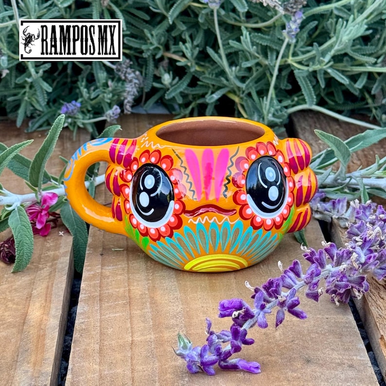 May include: Orange ceramic mug with a whimsical face design. The mug features large black eyes, a smiling mouth, and colorful floral patterns in red, pink, and turquoise. The handle is curved, and the mug is set against a wooden surface with lavender.