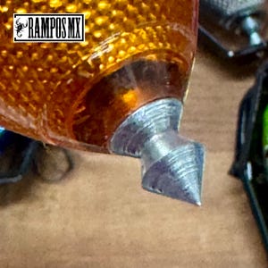 K&ouml;nnte beinhalten: Nahaufnahme eines metallischen silber-orangenen Objekts. Das Objekt hat ein spitzes Ende und eine strukturierte, orangefarbene, abgerundete Oberseite. Der Text "RAMPOS MX" ist in einem wei&szlig;en Feld oben links zu sehen.