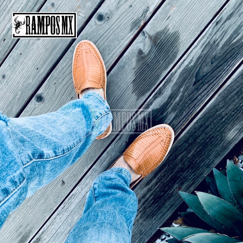 May include: A pair of tan leather sandals with woven straps. The sandals are being worn by a person wearing blue jeans. The sandals are on a wooden deck.