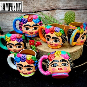 May include: Six colorful ceramic mugs with a Frida Kahlo inspired design. Each mug features a different color scheme and floral details. The mugs are painted with a whimsical style and have a unique handle.