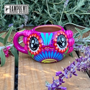 May include: A vibrant, hand-painted ceramic mug with a whimsical design. The mug features a bright pink base, a large handle, and is adorned with colorful floral and face-like patterns. The text "RAMPOS MX" is visible in the upper left corner.