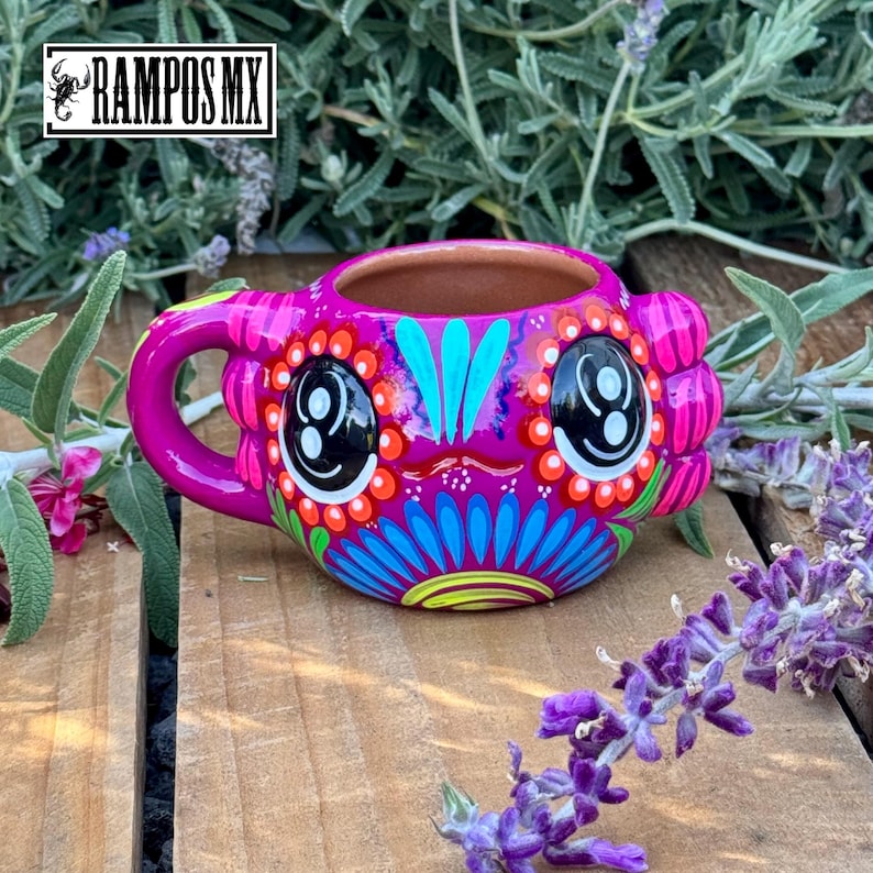 May include: A vibrant, hand-painted ceramic mug with a whimsical design. The mug features a bright pink base, a large handle, and is adorned with colorful floral and face-like patterns. The text "RAMPOS MX" is visible in the upper left corner.