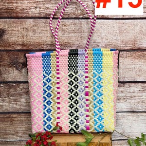 May include: A rectangular tote bag woven from plastic strips in a vertical striped pattern of pink, blue, black, lavender, gold, and yellow. The bag features two braided handles in pink and white, and includes the text RAMPOS MX and #15.