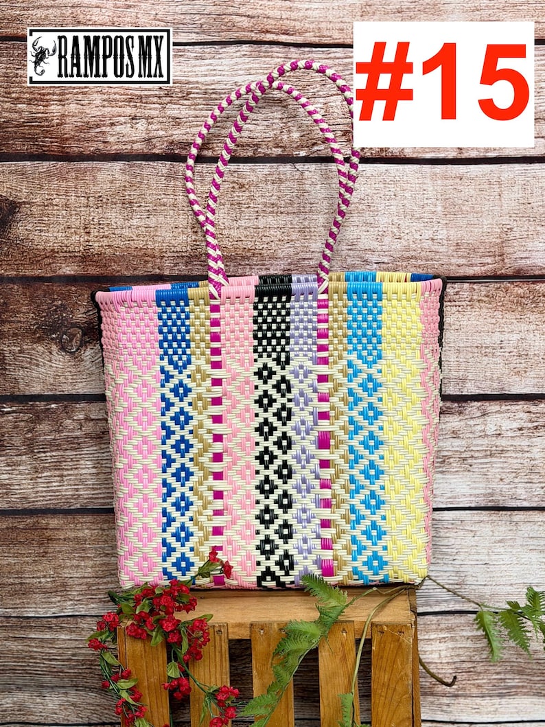 May include: A rectangular tote bag woven from plastic strips in a vertical striped pattern of pink, blue, black, lavender, gold, and yellow. The bag features two braided handles in pink and white, and includes the text RAMPOS MX and #15.