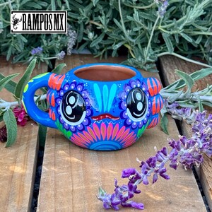 May include: A vibrant blue ceramic mug with a whimsical face design. The mug features large, stylized eyes, a red mouth, and colorful floral patterns in pink, orange, and purple. The handle is blue, and the interior is a natural terracotta color. The mug has the text "RAMPOS MX".
