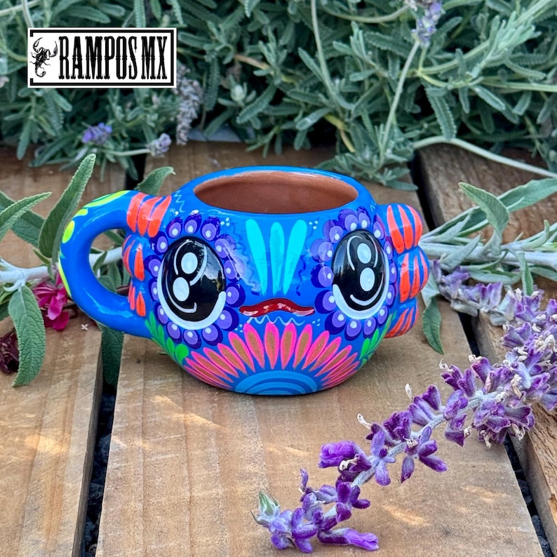 May include: A vibrant blue ceramic mug with a whimsical face design. The mug features large, stylized eyes, a red mouth, and colorful floral patterns in pink, orange, and purple. The handle is blue, and the interior is a natural terracotta color. The mug has the text "RAMPOS MX".