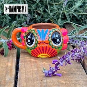 May include: A hand-painted ceramic mug with a vibrant, colorful design. The mug features an orange base with a face-like design, including large black eyes, and is accented with pink, blue, and gold details. The handle is orange. The mug is on a wooden surface.
