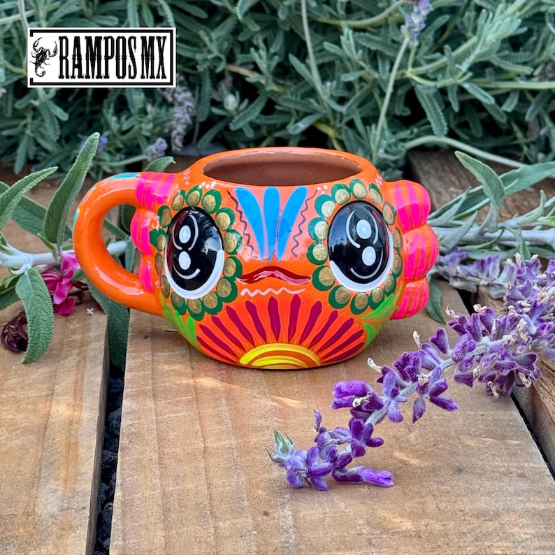May include: A hand-painted ceramic mug with a vibrant, colorful design. The mug features an orange base with a face-like design, including large black eyes, and is accented with pink, blue, and gold details. The handle is orange. The mug is on a wooden surface.