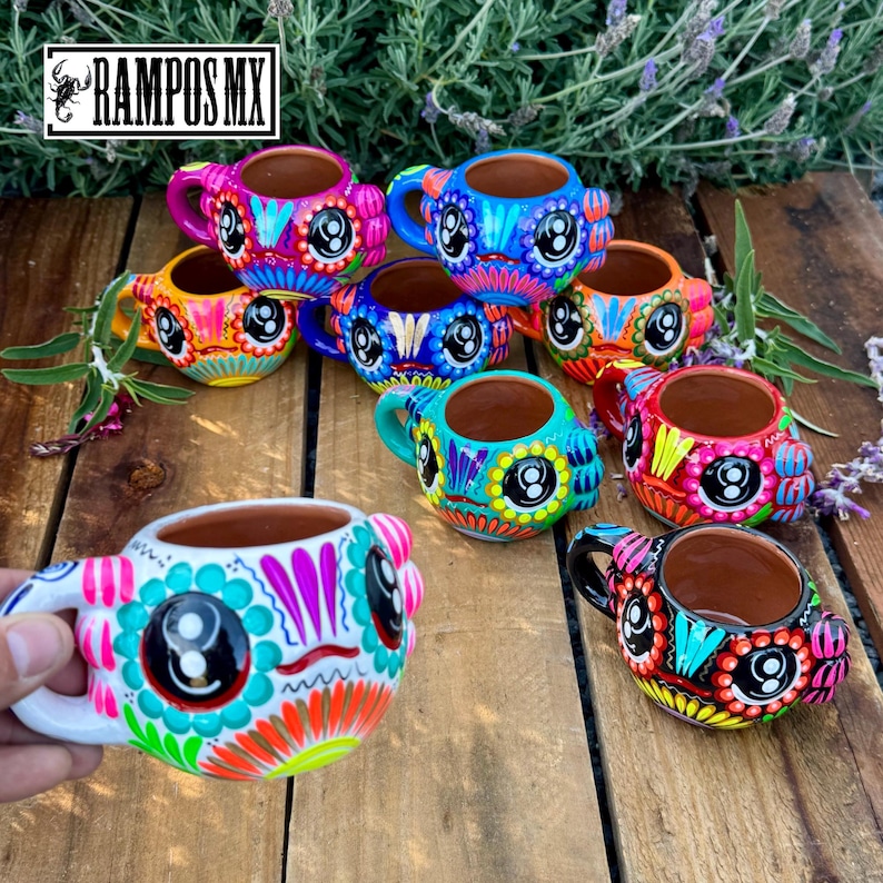 May include: A collection of colorful, hand-painted ceramic mugs with a Day of the Dead sugar skull design. Each mug features vibrant colors, including pink, blue, orange, and green, with intricate patterns and a handle. The mugs are displayed on a wooden surface.