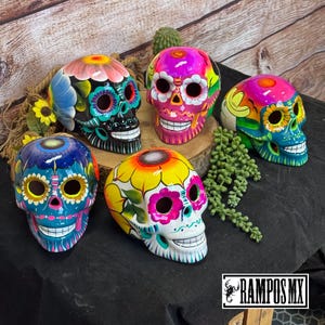 May include: A collection of six hand-painted ceramic sugar skulls, each with unique floral designs and vibrant colors. The skulls feature bright hues of pink, blue, yellow, and green, with intricate details and smiling teeth. The skulls are displayed on a wooden surface.