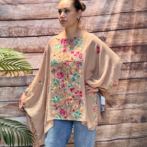 Könnte beinhalten: Ein beiges Oberteil im Poncho-Stil mit lebendiger, mehrfarbiger Blumen- und Schmetterlingsstickerei auf der Vorderseite und den Ärmeln. Das Kleidungsstück hat eine lockere, fließende Silhouette mit weiten Ärmeln und wird mit einer hellblauen Jeans kombiniert.