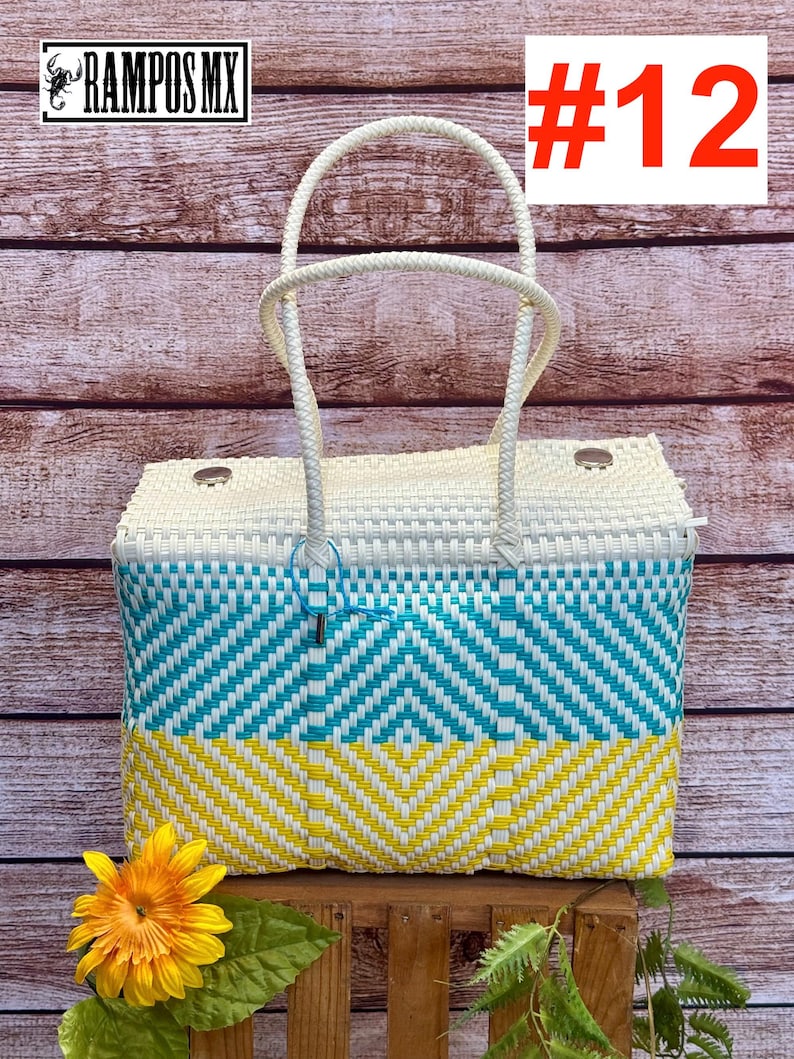 Op de afbeelding: Een rechthoekige gevlochten draagtas met een wit bovengedeelte, een turquoise middenband en een geel ondergedeelte, allemaal voorzien van een chevronpatroon. De tas heeft twee witte handvatten en twee zilverkleurige metalen noppen op de deksel.