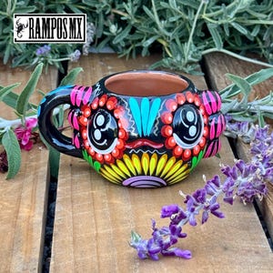May include: A hand-painted ceramic mug with a black handle and exterior. The mug features a colorful Day of the Dead design with red, yellow, pink, and turquoise accents. The mug has the text "RAMPOS MX" in a white box.