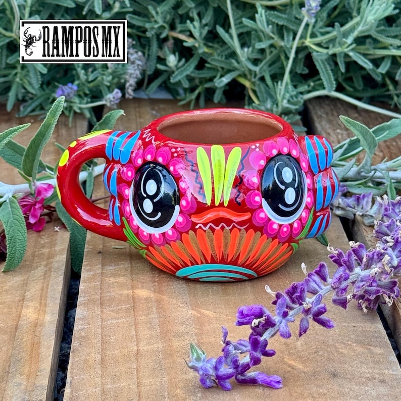 May include: A vibrant, hand-painted ceramic mug with a whimsical design. The mug features a red base with colorful floral and geometric patterns, including large eye-like shapes. The handle and rim are also red. The mug has the text "RAMPOS MX".