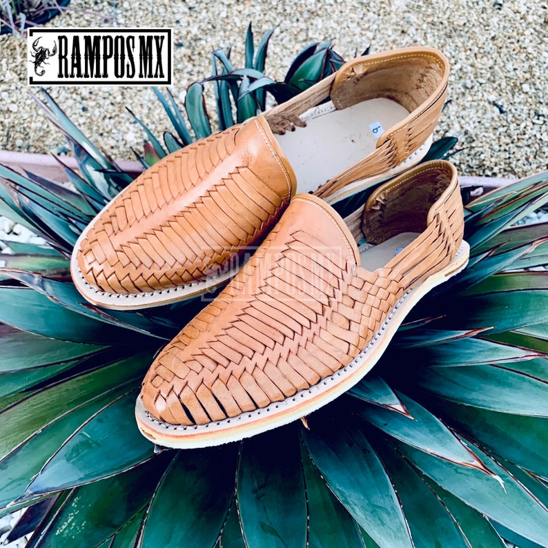 May include: A pair of tan leather sandals with woven straps. The sandals are on a green leafy background.