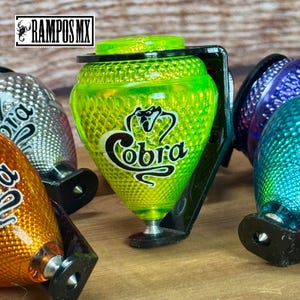 K&ouml;nnte beinhalten: Eine Sammlung bunter Kreisel. Der Hauptkreisel ist leuchtend gr&uuml;n mit dem Wort "Cobra" in wei&szlig;er Schrift. Die Kreisel haben eine strukturierte Oberfl&auml;che und sind auf schwarzen Halterungen montiert. Das Logo "RAMPOS MX" befindet sich oben links.