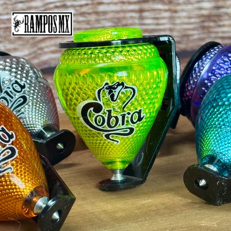 K&ouml;nnte beinhalten: Eine Sammlung bunter Kreisel. Der Hauptkreisel ist leuchtend gr&uuml;n mit dem Wort "Cobra" in wei&szlig;er Schrift. Die Kreisel haben eine strukturierte Oberfl&auml;che und sind auf schwarzen Halterungen montiert. Das Logo "RAMPOS MX" befindet sich oben links.