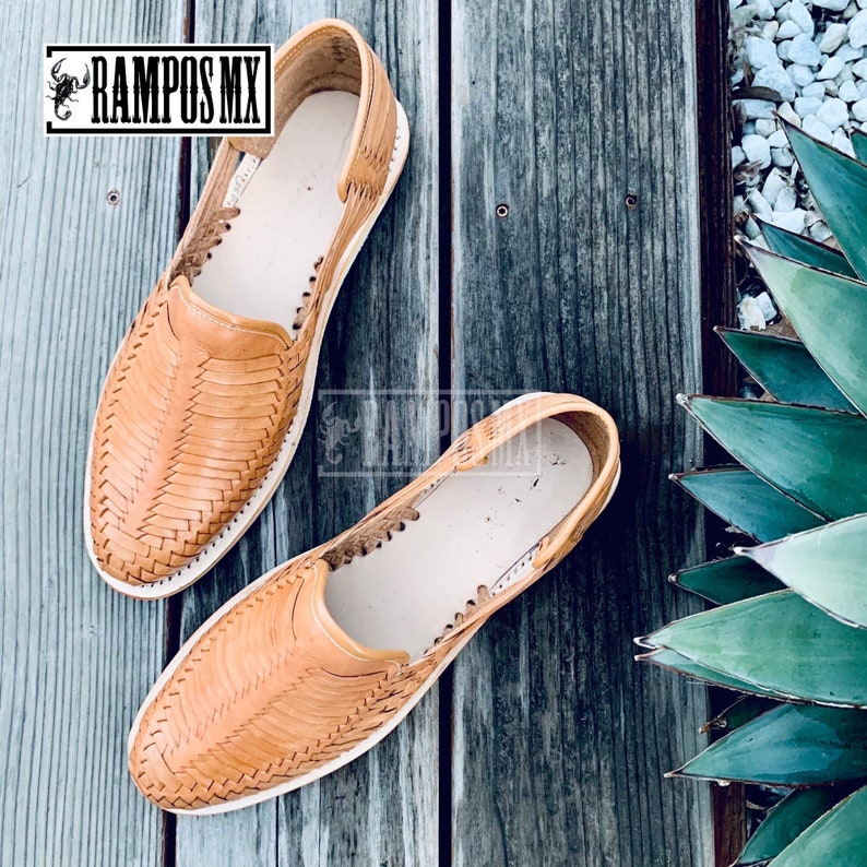 May include: A pair of tan leather sandals with a woven design. The sandals are on a wooden surface.