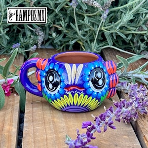 May include: A vibrantly painted ceramic mug with a whimsical design. The mug features a blue base with large, stylized eyes and colorful accents of orange, yellow, and purple. The handle is a matching blue. The text "RAMPOS MX" is in the upper left corner.