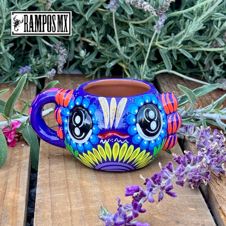 May include: A vibrantly painted ceramic mug with a whimsical design. The mug features a blue base with large, stylized eyes and colorful accents of orange, yellow, and purple. The handle is a matching blue. The text "RAMPOS MX" is in the upper left corner.