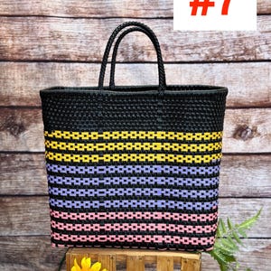 May include: A black woven tote bag with yellow, lavender, and pink horizontal stripes. The bag has two black handles and is displayed on a wooden crate. The number 7 is displayed in red on a white background.