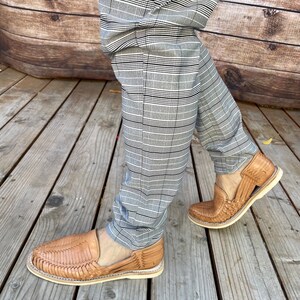May include: A pair of tan leather sandals with a woven design. The sandals are being worn by a person wearing grey and black checked trousers. The person is standing on a wooden deck.
