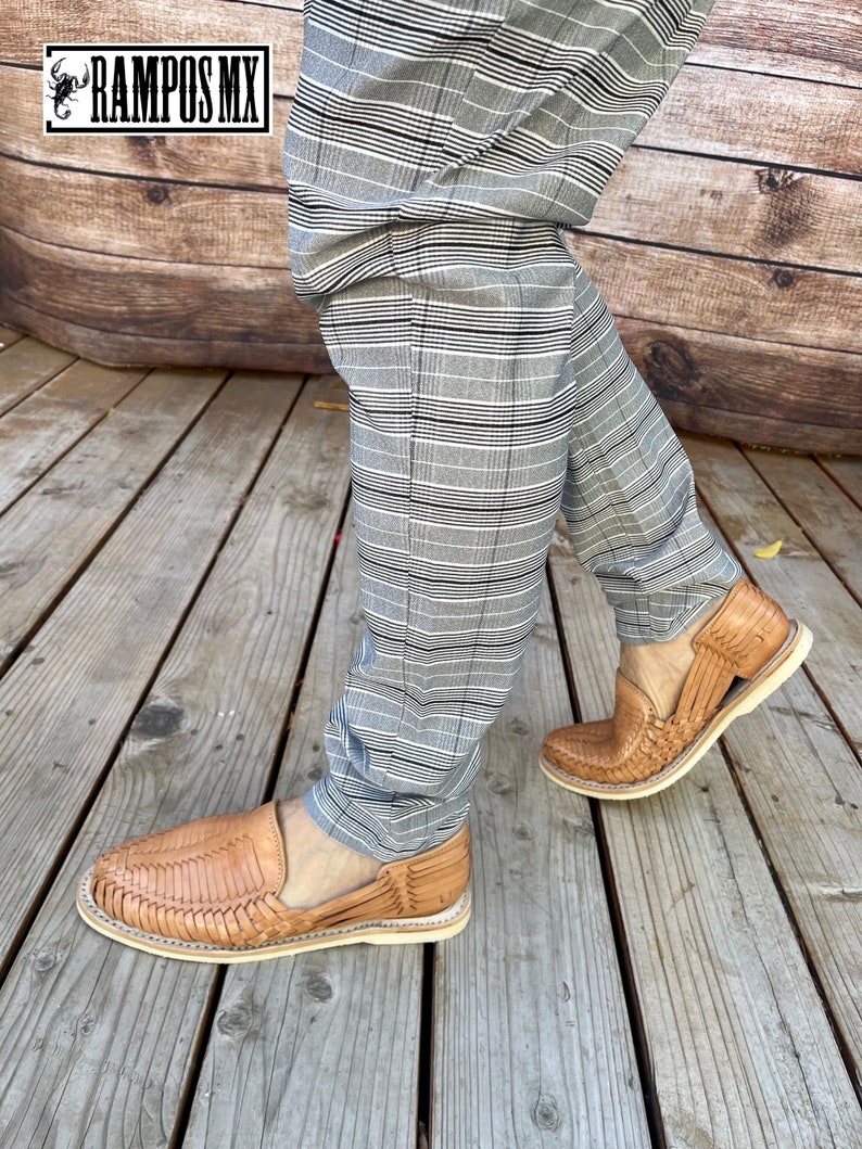 May include: A pair of tan leather sandals with a woven design. The sandals are being worn by a person wearing grey and black checked trousers. The person is standing on a wooden deck.