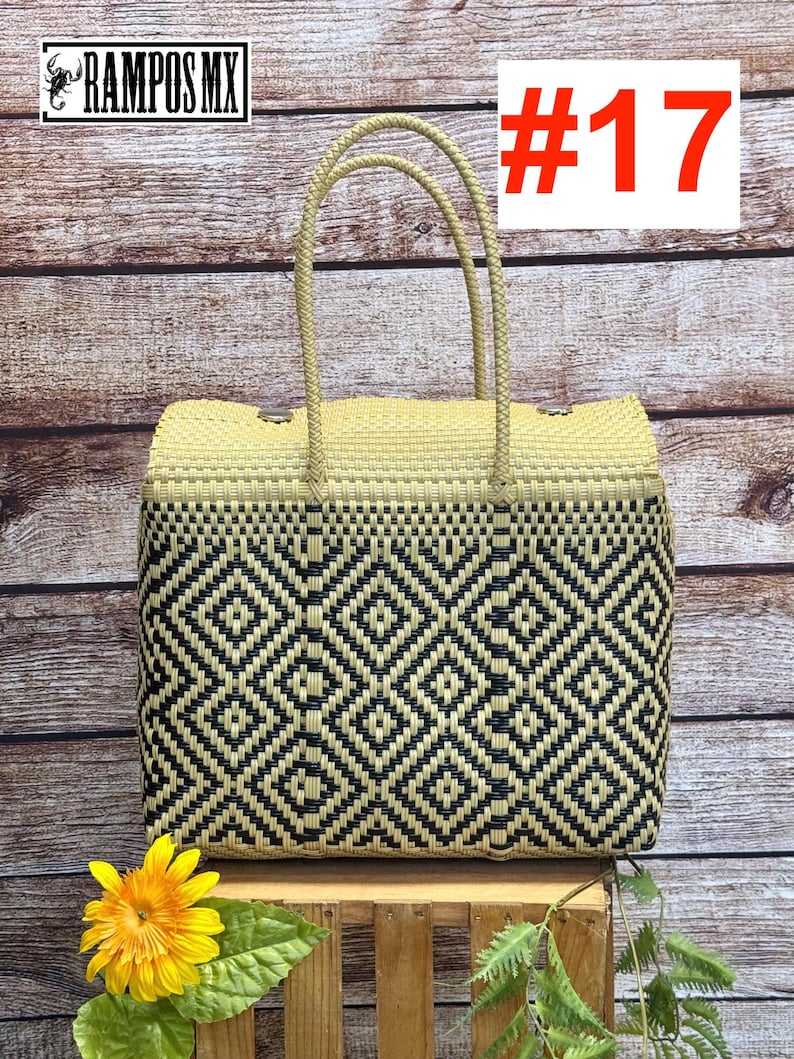 Op de afbeelding: Een rechthoekige draagtas met een geweven patroon van zwarte en beige plastic strips. De tas heeft twee bijpassende beige gevlochten handvatten en een bovenflap die is vastgezet met zilverkleurige metalen knopen. De tekst RAMPOS MX en #17 is zichtbaar.