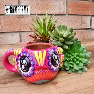 May include: A vibrant pink ceramic mug with a whimsical face design, featuring large black eyes, yellow accents, and purple floral details. The mug is decorated with a colorful, hand-painted design. The mug is displayed with green succulents on a wooden surface.