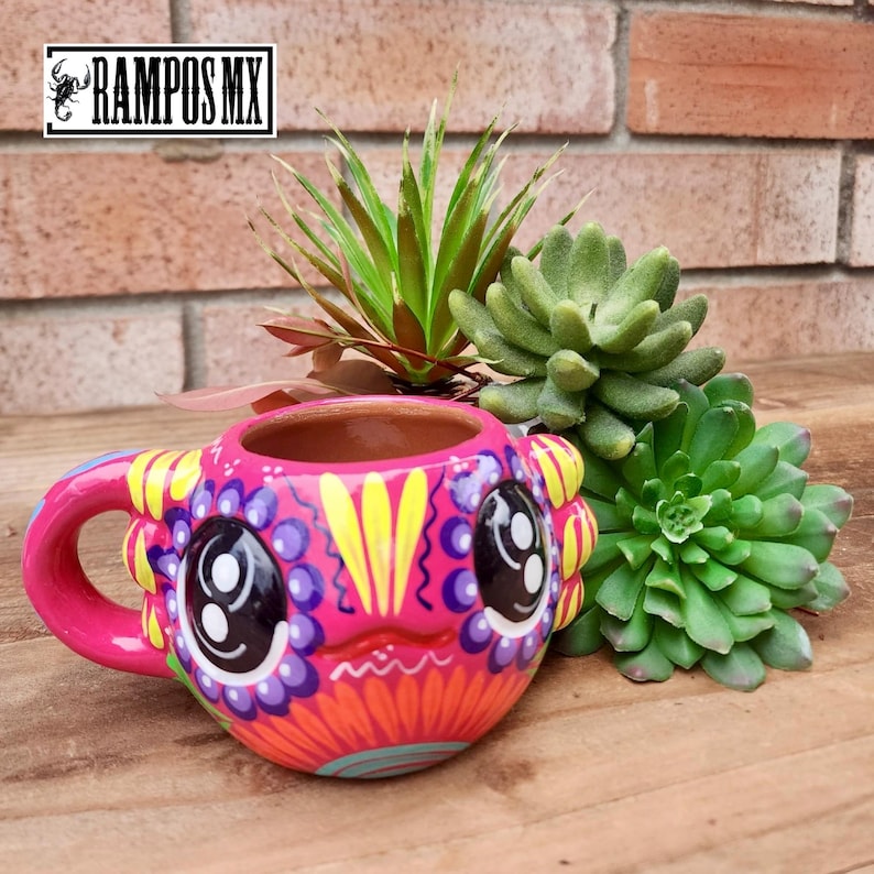 May include: A vibrant pink ceramic mug with a whimsical face design, featuring large black eyes, yellow accents, and purple floral details. The mug is decorated with a colorful, hand-painted design. The mug is displayed with green succulents on a wooden surface.