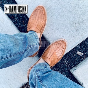 May include: A pair of tan leather sandals with a woven design. The sandals are being worn by someone wearing blue jeans.