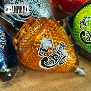 K&ouml;nnte beinhalten: Ein orangefarbener Kreisel mit strukturierter Oberfl&auml;che und dem Wort "Cobra" in wei&szlig;er Schrift. Der Kreisel hat eine silberne Spitze und ein Schlangendesign. Weitere Kreisel in verschiedenen Farben sind im Hintergrund zu sehen.