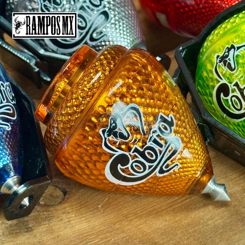 K&ouml;nnte beinhalten: Ein orangefarbener Kreisel mit strukturierter Oberfl&auml;che und dem Wort "Cobra" in wei&szlig;er Schrift. Der Kreisel hat eine silberne Spitze und ein Schlangendesign. Weitere Kreisel in verschiedenen Farben sind im Hintergrund zu sehen.