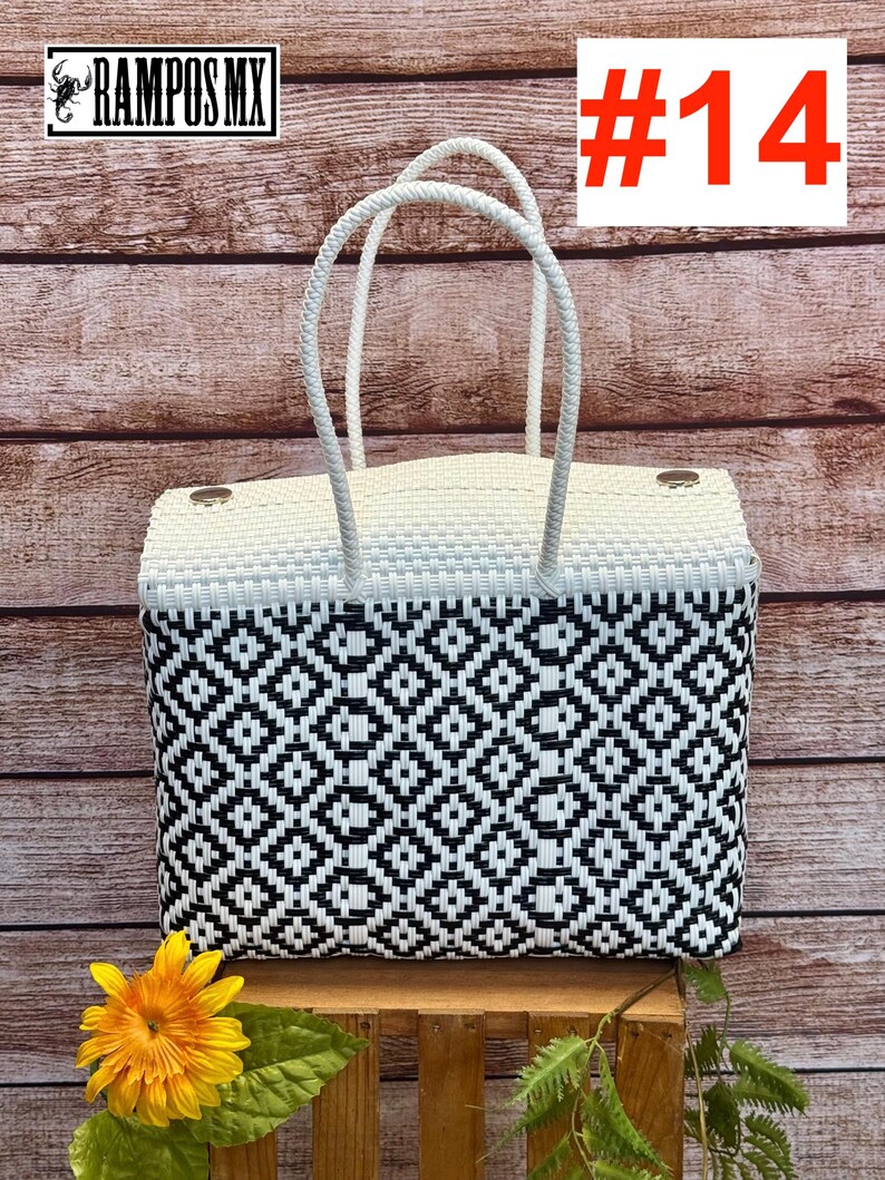 Op de afbeelding: Een rechthoekige gevlochten draagtas met een zwart-wit geometrisch ruitpatroon op het lichaam en een effen wit bovengedeelte. De tas heeft twee witte gevlochten handvatten en twee zilverkleurige metalen sluitingen. Tekst RAMPOS MX #14.