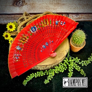 May include: A vibrant red hand fan with floral designs in blue, yellow, and green. The fan is open, displaying its intricate painted details. The image also includes a small cactus in a terracotta pot and the text "RAMPOS MX".