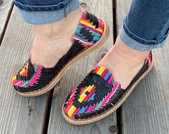 Puede incluir: Un par de sandalias de cuero negro con correas tejidas de colores. Las sandalias tienen una suela plana y están hechas en un estilo mexicano tradicional.