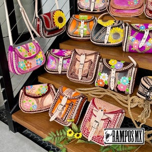 May include: A selection of handcrafted leather crossbody bags in various colours, including pink, orange, and brown. Each bag features floral designs, intricate stitching, and a buckle closure. The bags are displayed on wooden steps.