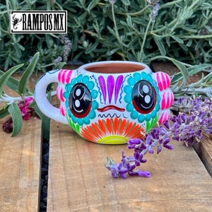 May include: A hand-painted ceramic mug with a colorful Day of the Dead design. The mug features a white base with a face design in teal, pink, orange, and black. The handle is white, and the mug is sitting on a wooden surface.