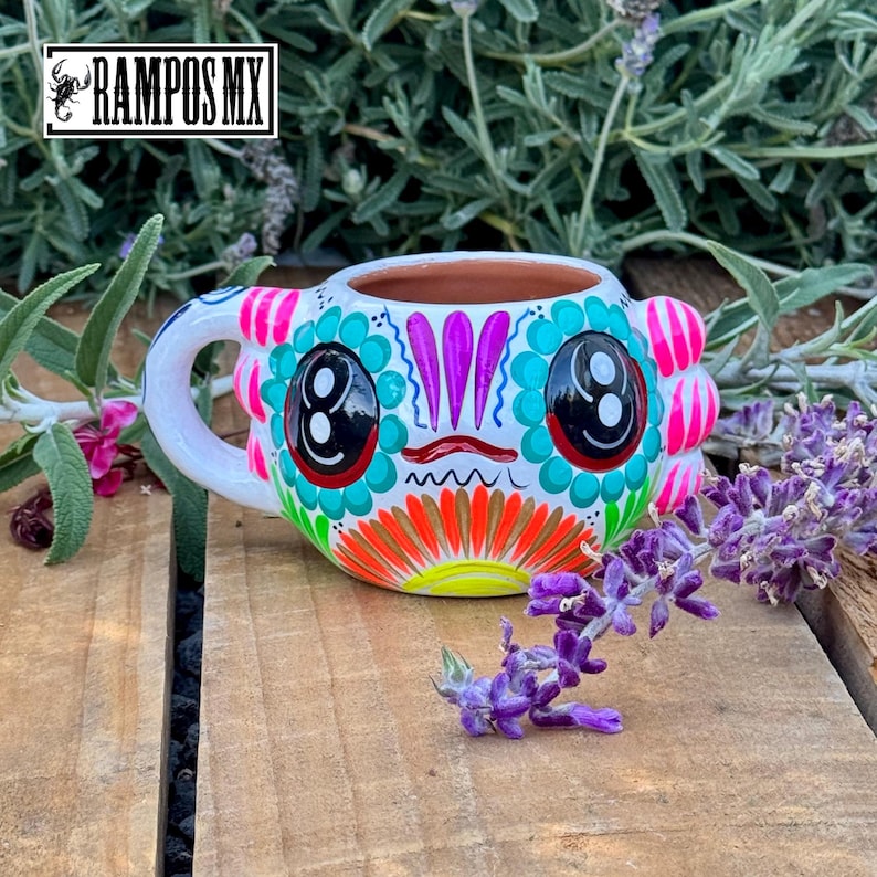 May include: A hand-painted ceramic mug with a colorful Day of the Dead design. The mug features a white base with a face design in teal, pink, orange, and black. The handle is white, and the mug is sitting on a wooden surface.