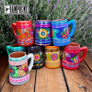 May include: A collection of vibrantly painted ceramic mugs with handles. Each mug features a unique design with floral and bird motifs in red, blue, green, orange, and yellow. The mugs are displayed on a wooden surface with a lavender background. The text "RAMPOS MX" is visible.