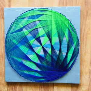 May include: A blue and green string art design in the shape of a circle. The design is made up of many intersecting lines, creating a geometric pattern.