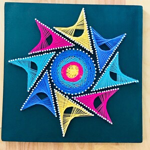 May include: A colorful string art piece with a starburst design. The starburst is made of yellow, blue, and pink string wrapped around nails on a dark green background.
