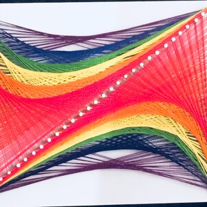 May include: A rainbow string art piece with a white background. The string is wrapped around nails in a curved pattern, creating a colorful and abstract design.