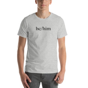 Pode incluir: Uma camiseta cinza mescla com o texto "he/him" impresso em preto na frente.
