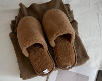 Può includere: Un paio di pantofole marroni e pelose con un'etichetta di taglia che recita "36-37". Le pantofole sono sopra un maglione marrone piegato.