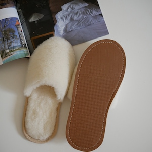 Pu&ograve; includere: Un paio di pantofole in pelle di pecora bianca con suole in pelle marrone. Le pantofole sono disposte su una superficie bianca.