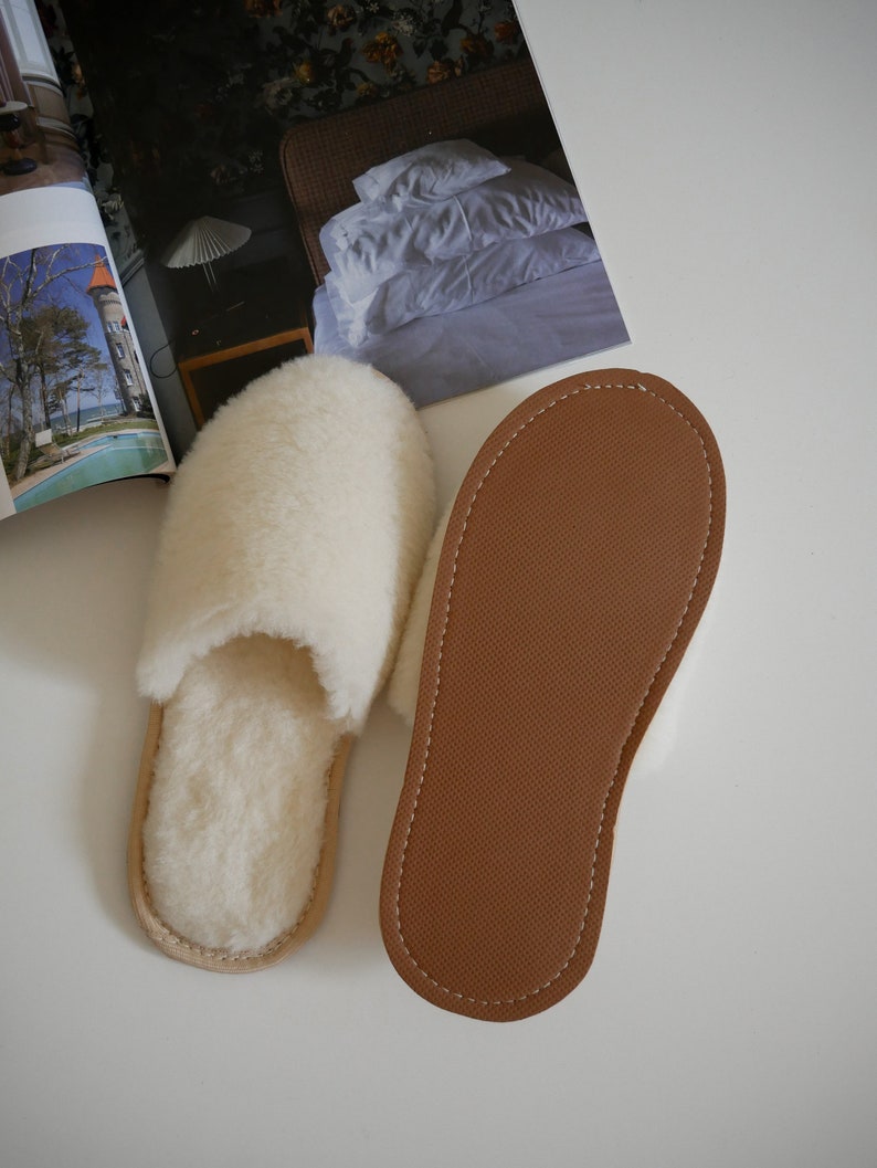 Pu&ograve; includere: Un paio di pantofole in pelle di pecora bianca con suole in pelle marrone. Le pantofole sono disposte su una superficie bianca.