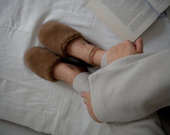 May include: A pair of brown fuzzy slippers with a white background. The slippers are worn by a person wearing a white sweater.