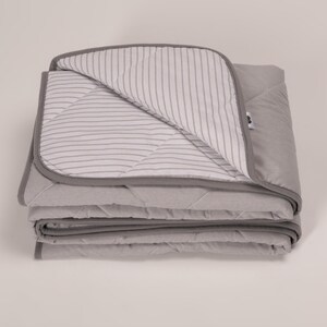 May include: A gray quilted blanket with a white and gray striped lining. The blanket is folded in thirds and is lying on a white surface.
